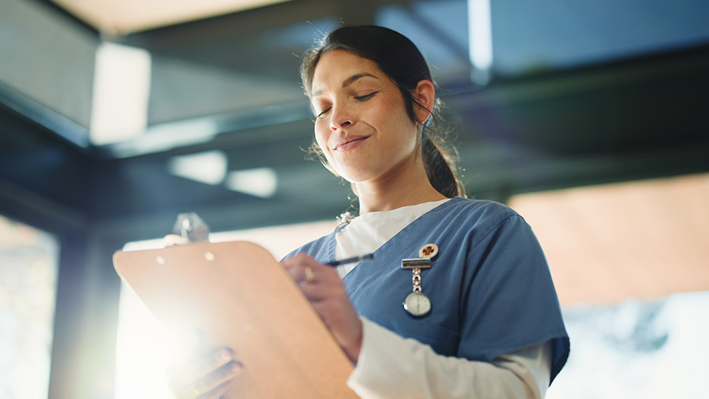 A nurse in blue scrubs smiling, holding a clipboard, sunlight streaming in. The scene conveys professionalism and warmth in a healthcare setting.