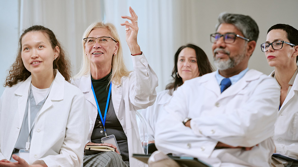 Group of medical professionals in white lab coats sitting attentively. A blonde woman raises her hand to speak, suggesting a seminar or discussion setting.