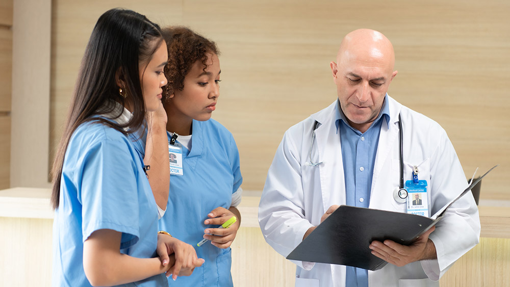 A doctor in a white coat reviews a clipboard with two medical students in blue scrubs. They appear focused and engaged in a hospital setting.